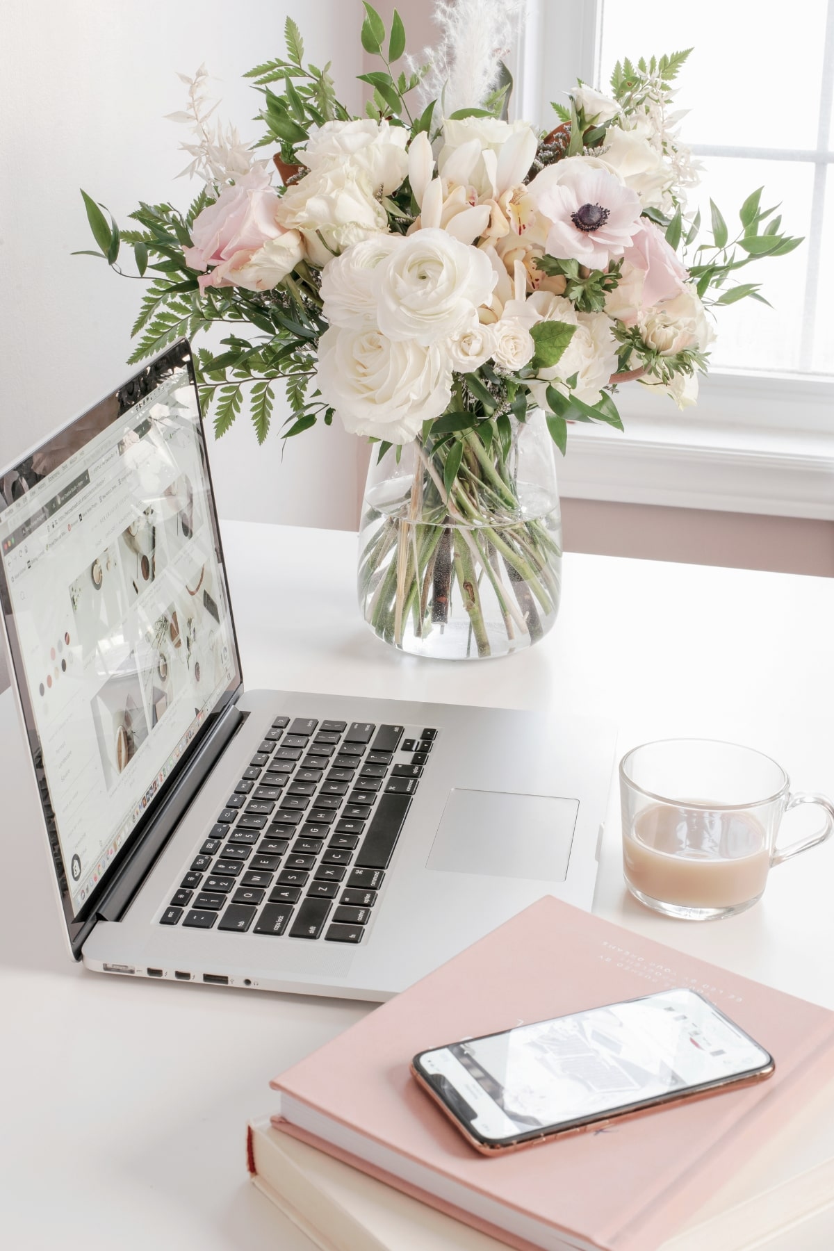 白とピンクを基調としたデスク上に、ノートパソコンとスマートフォン、花瓶に生けられた白い花束が置かれた画像 - ホームページ制作でのお悩み